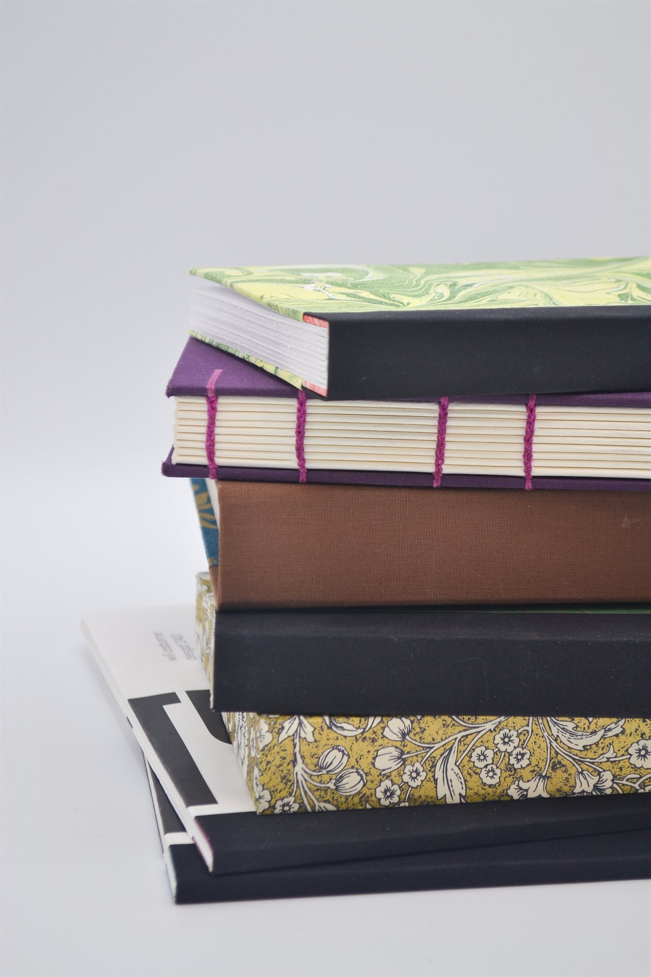 A stack of bound books viewed from the spine edge, topped with a green marbled cover and black cloth spine, above a purple book with exposed pink stitching, a brown cloth spine, a black spine, and a book covered in yellow floral patterned paper