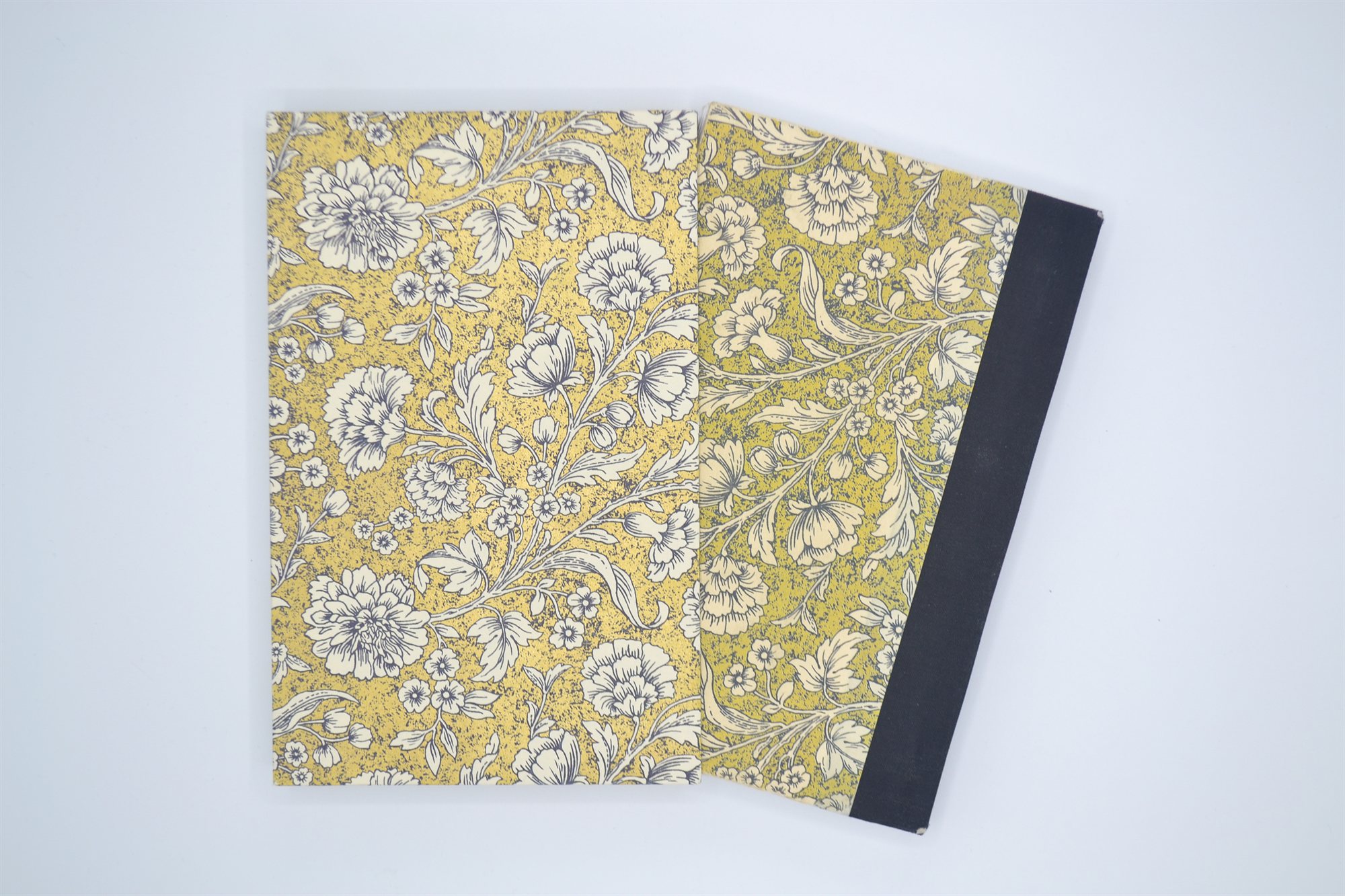 Two books lying flat side by side, both covered in matching cream and gold floral patterned paper, one fitted with a black cloth spine