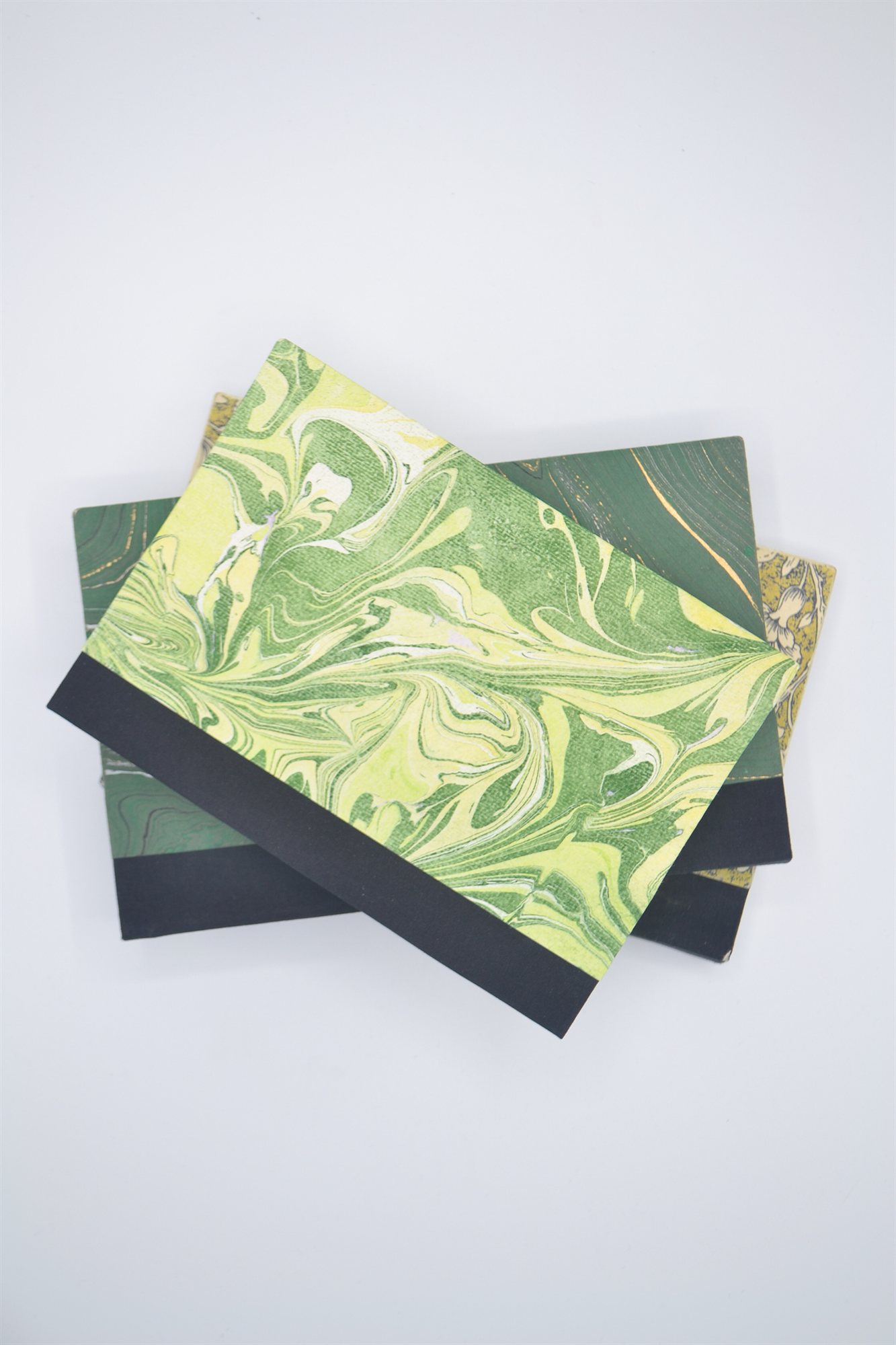Two books with green and yellow marbled paper covers and black cloth spines, overlapping on top of a stack of green and gold patterned books