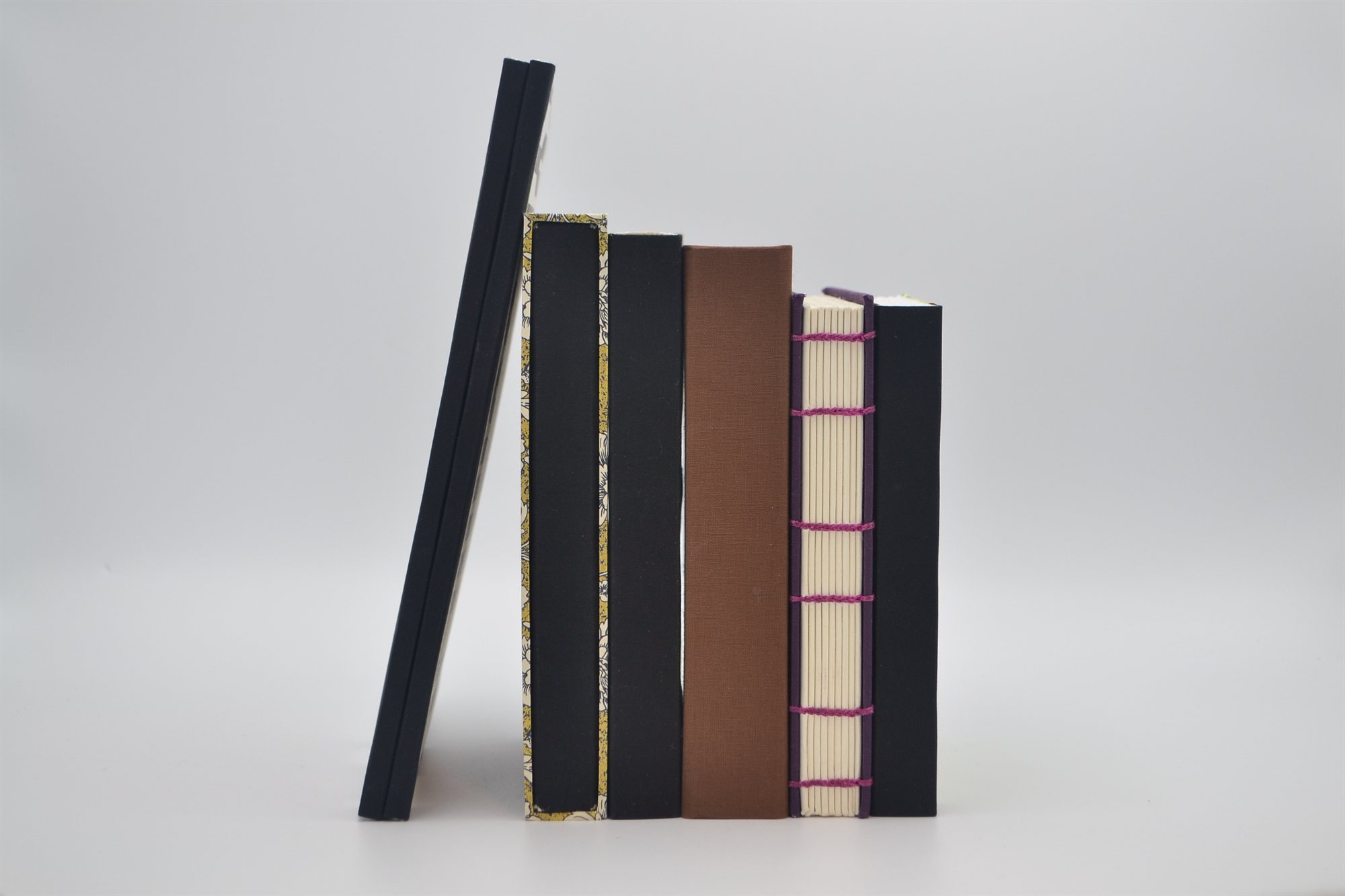 A row of finished books standing upright, showing black cloth, a floral pattern bordered in black, brown cloth, and purple cloth with exposed pink hand-sewn stitching along the spine
