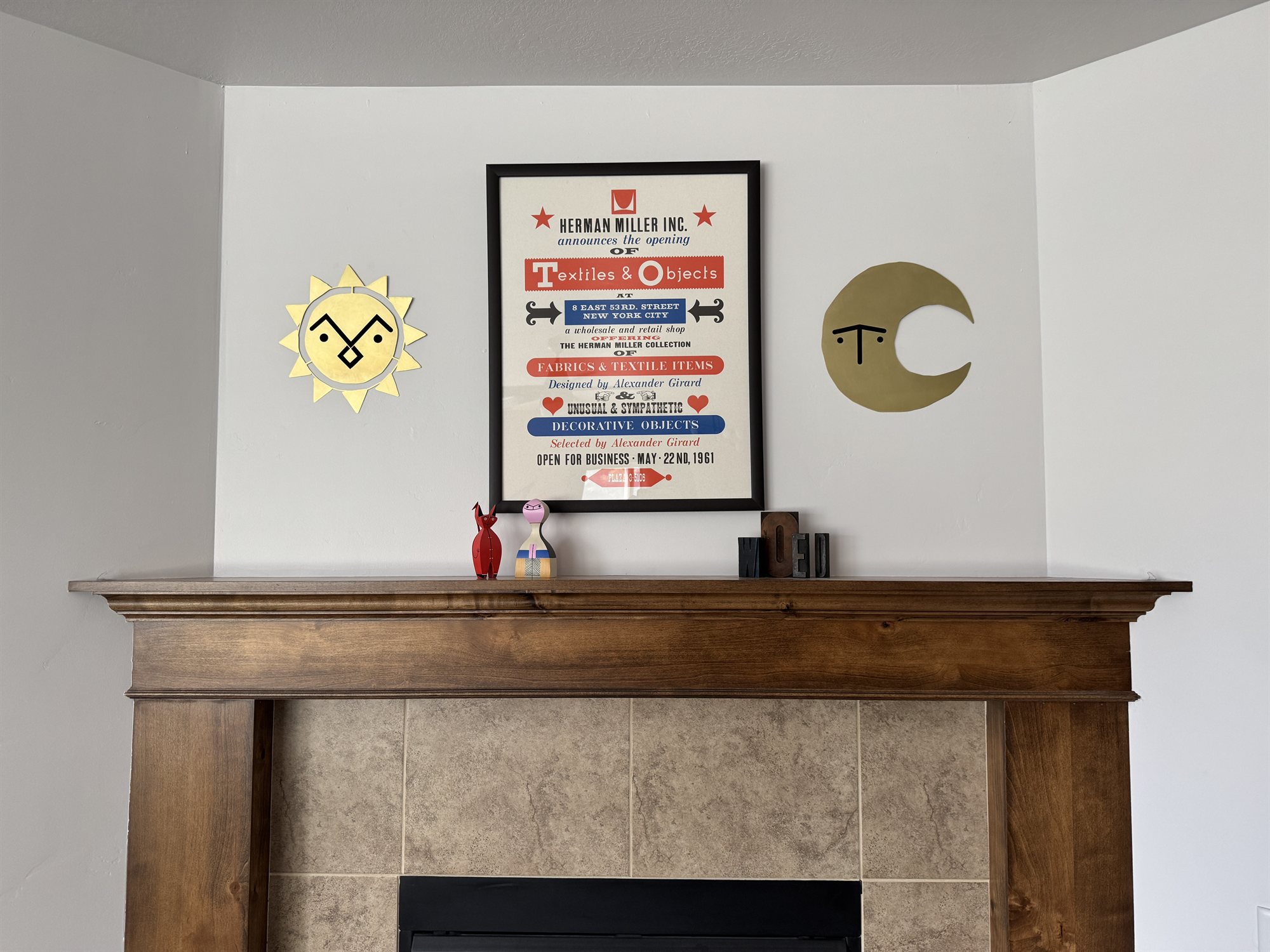 A polished brass sun and crescent moon, each with a grumpy cutout face, mounted on the wall flanking a framed vintage Herman Miller Textiles & Objects poster above a wood mantel