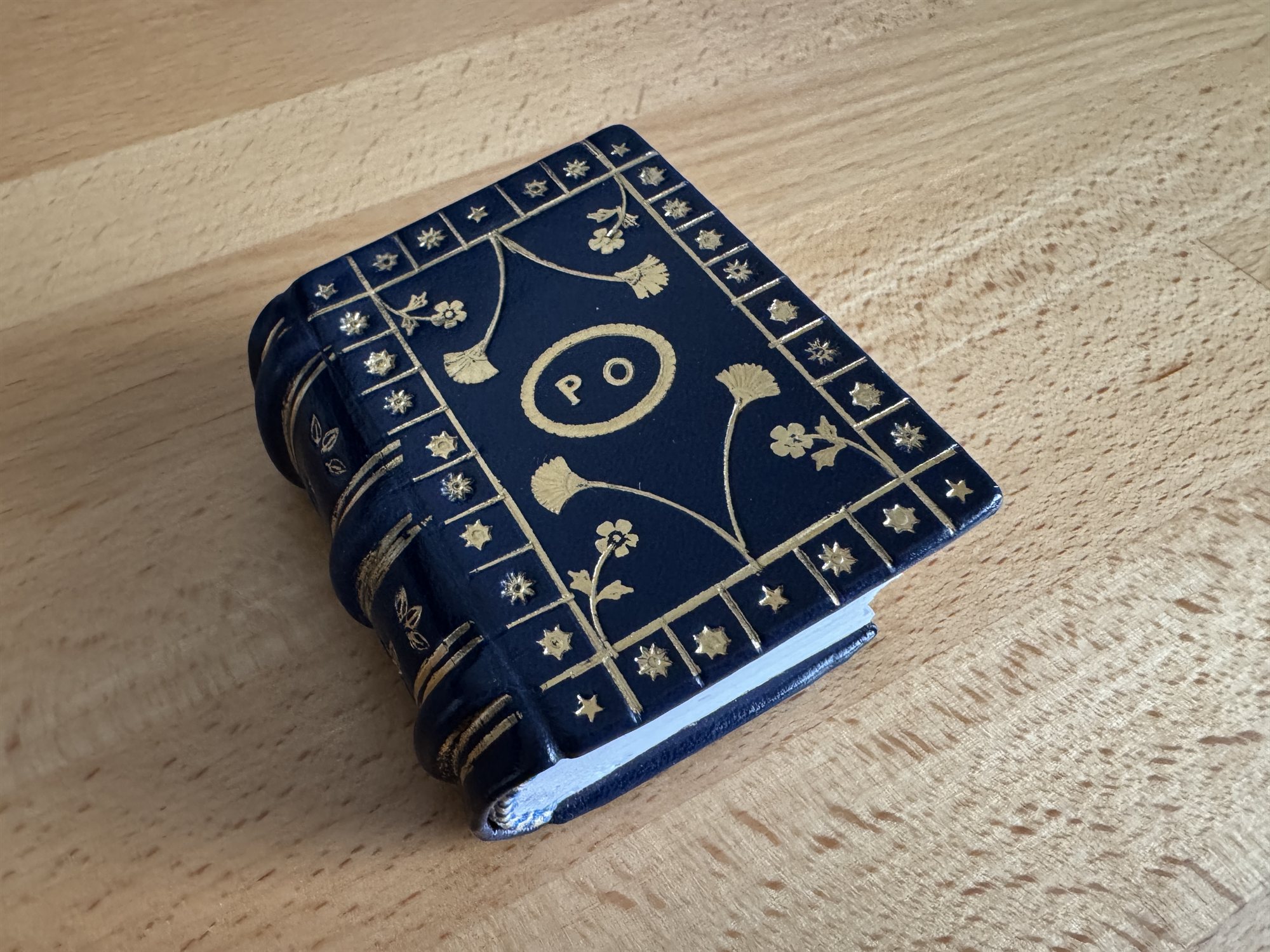 A miniature dark navy leather book with a gold-tooled floral border and an oval PO monogram stamped on the cover, representing the Fine Bookbinding project