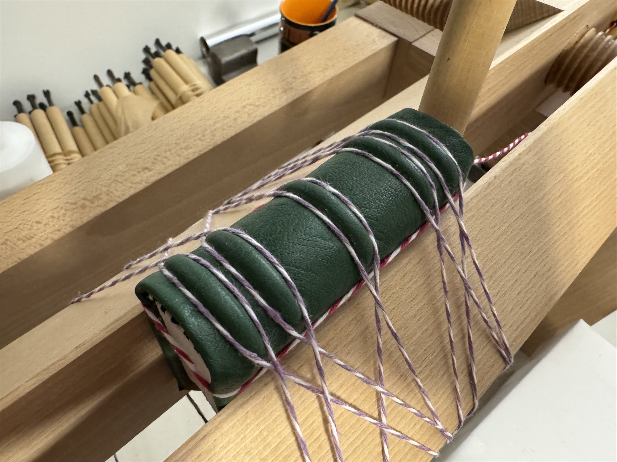 A green leather spine being laced onto its boards with cord on a wooden finishing press, the cords crossing in an X pattern