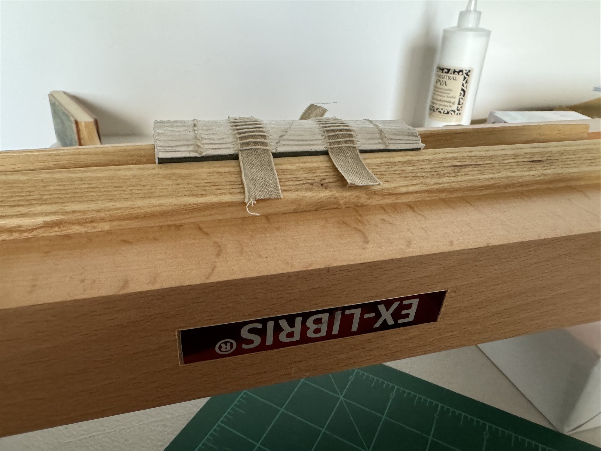 A sewn text block with cloth tapes resting on the workbench, glue and a bookplate box visible in the background