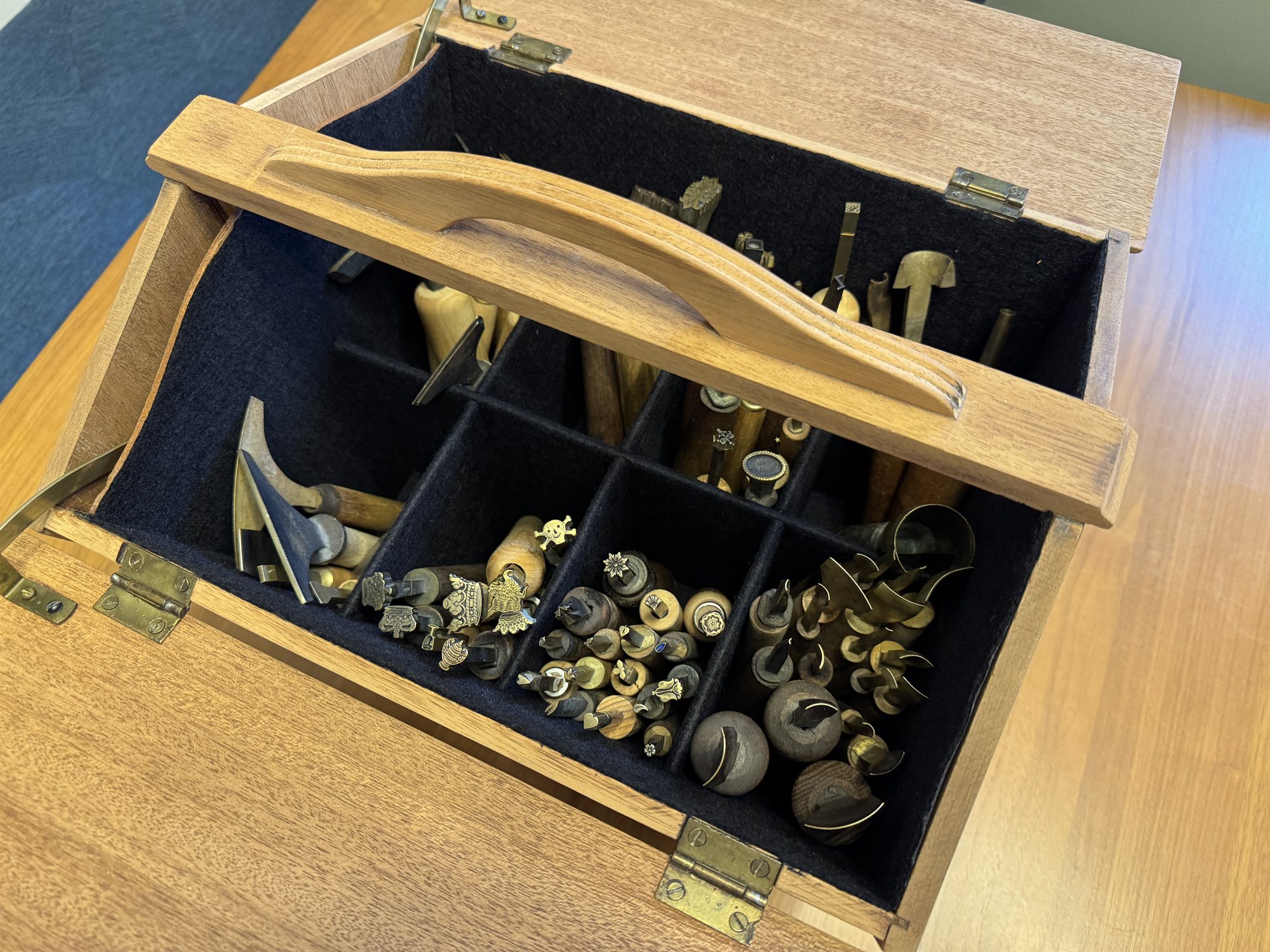 The open antique sewing box, refinished and fitted with felt-lined compartments holding rows of brass finishing stamps and hammers