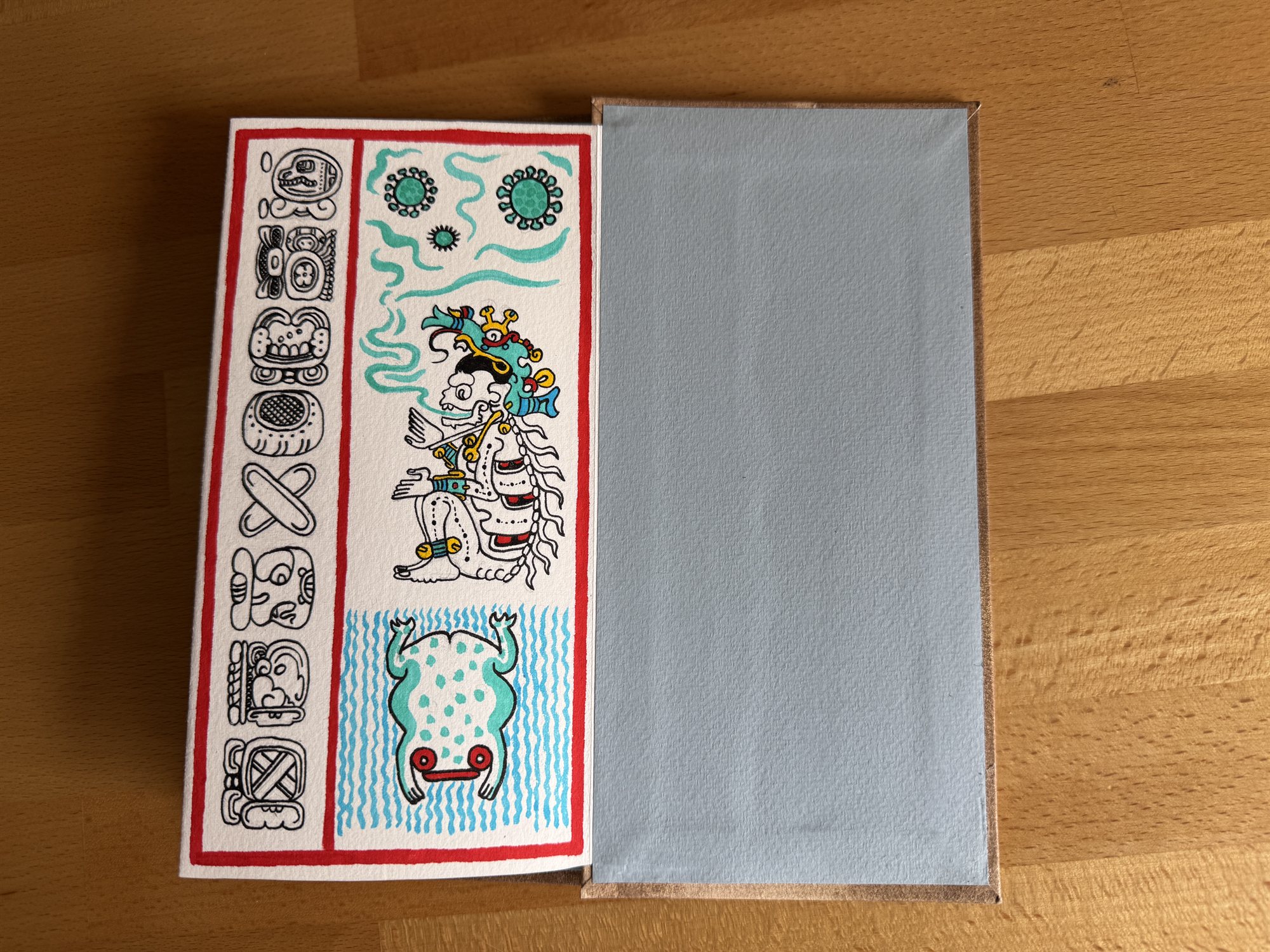 A page bordered in red showing a column of small glyphs beside a seated figure in a teal, yellow, and red headdress wreathed in smoke, with a spotted toad on wavy blue lines below, next to the book's plain pale blue-gray endpaper