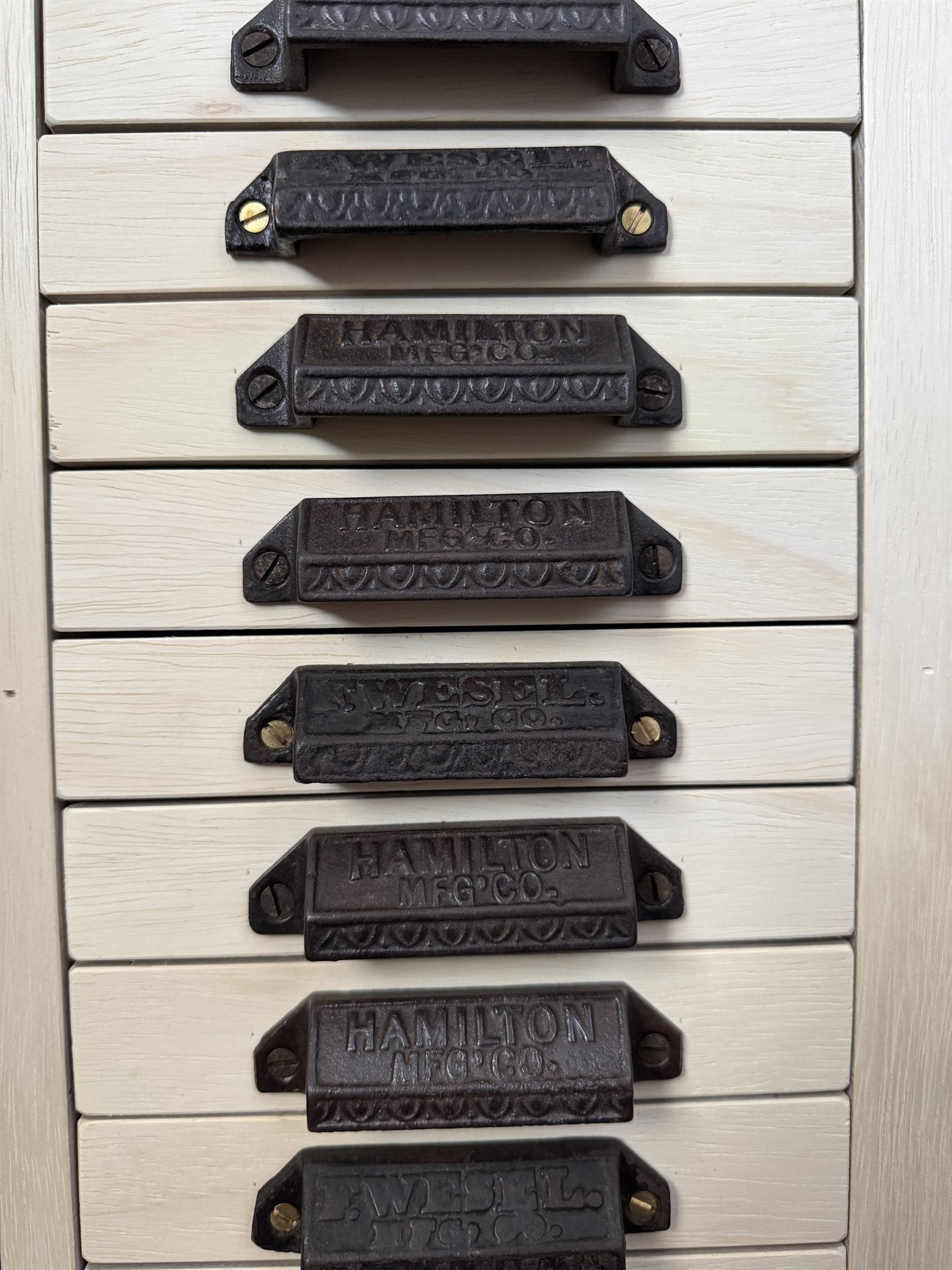 Close-up of the cast iron case pulls, alternating Hamilton Mfg Co and Wesel Mfg Co markings, screwed into the pale wood drawer fronts
