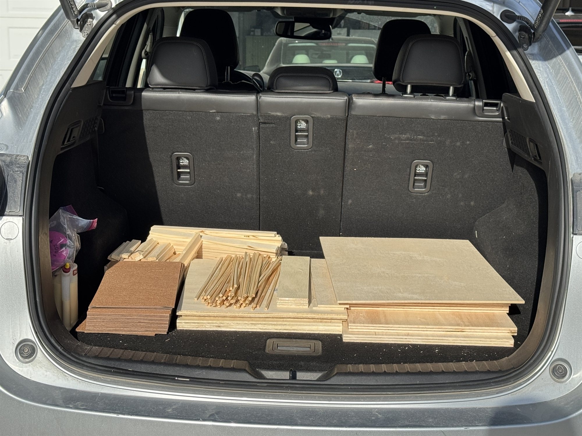 A car trunk loaded with cut sheets of plywood and particle board plus bundles of thin divider strips, the raw materials hauled home for the build
